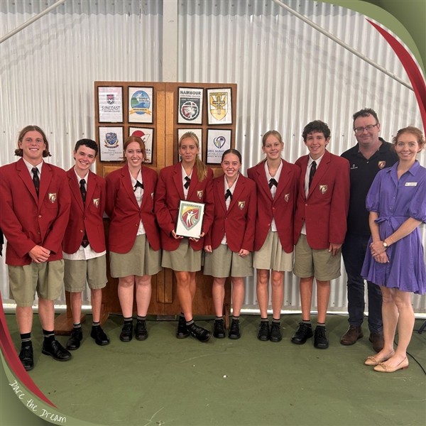 Students in red blazers standing together indoors, holding a framed school emblem, with staff members beside them.